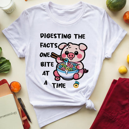 White t-shirt with a cartoon pig eating cereal and text, surrounded by vegetables and a glass of milk on a beige background.
