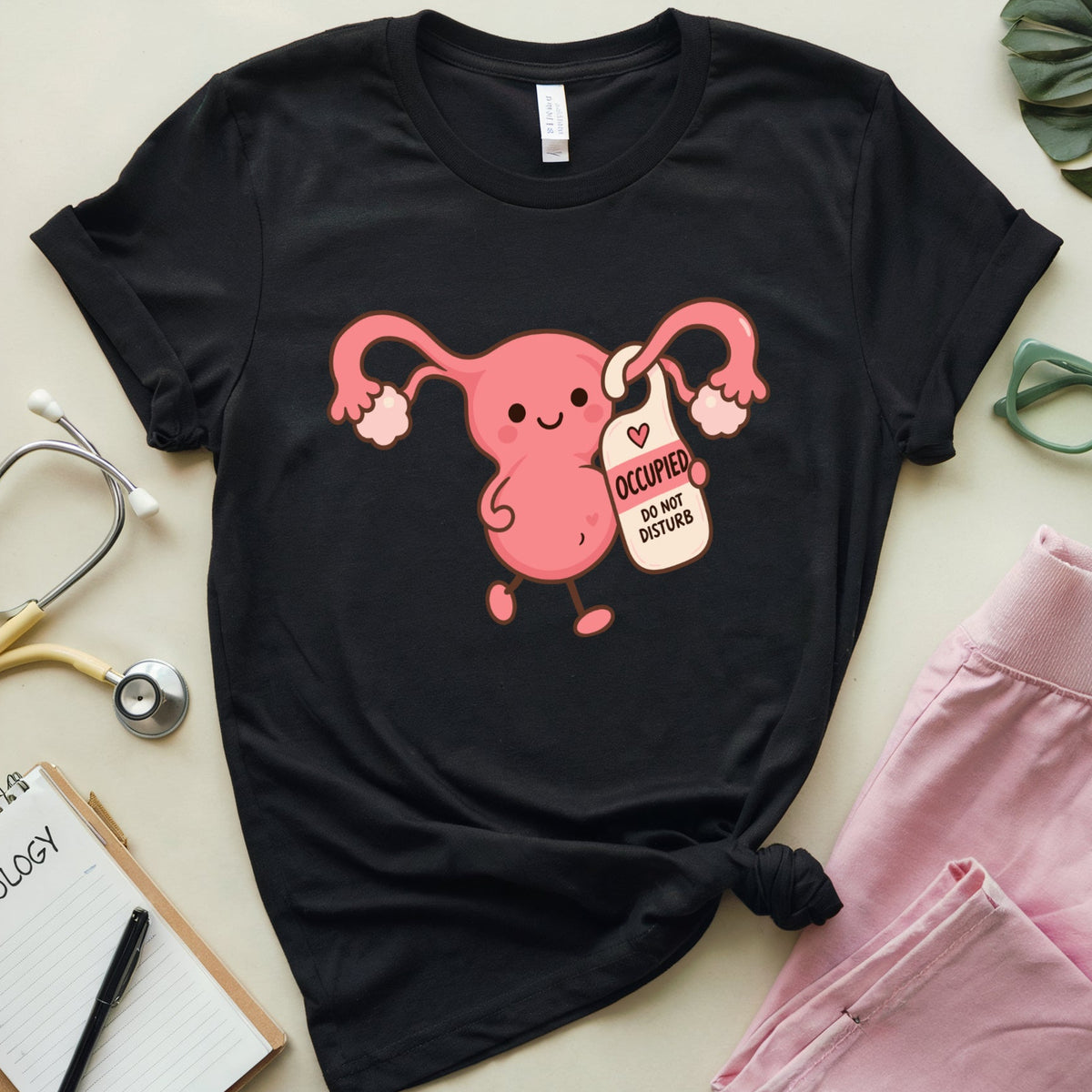 A black graphic t-shirt featuring a pink cartoon uterus holding a tag that reads 'OOCURRED OVARY DISTURBS' in a flat-lay arrangement with pink pants, a stethoscope, a notebook, and green eyeglasses on a light beige background.