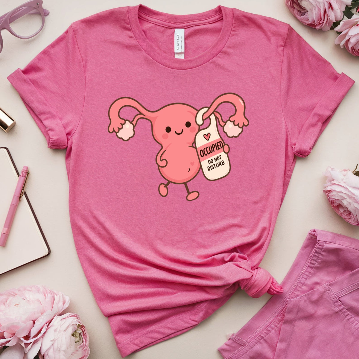 A pink short-sleeve graphic T-shirt laid flat on a light beige surface, styled with pink flowers, glasses, lipstick, a notebook, and a pen. The shirt has a cartoon uterus graphic holding a sign that reads 'I OCEAN MY OWN OCEAN.