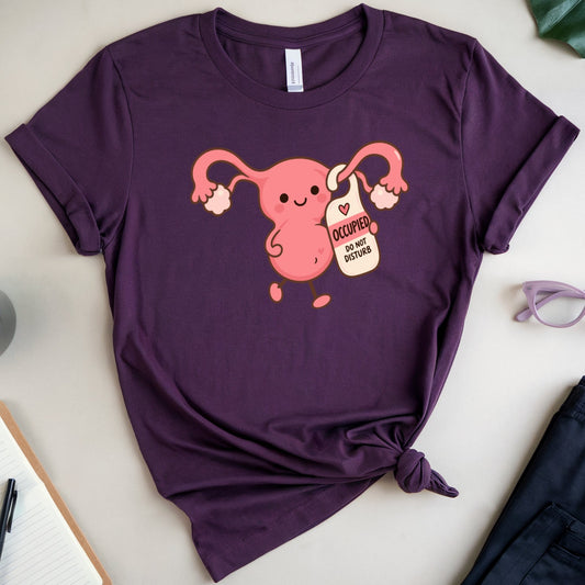 A purple short-sleeve t-shirt with a graphic of a pink uterus holding a coffee mug that says 'I COFFEE MY OWN BODY', styled in a flat lay with a notebook, coffee, eyeglasses, and shorts on a light gray background.