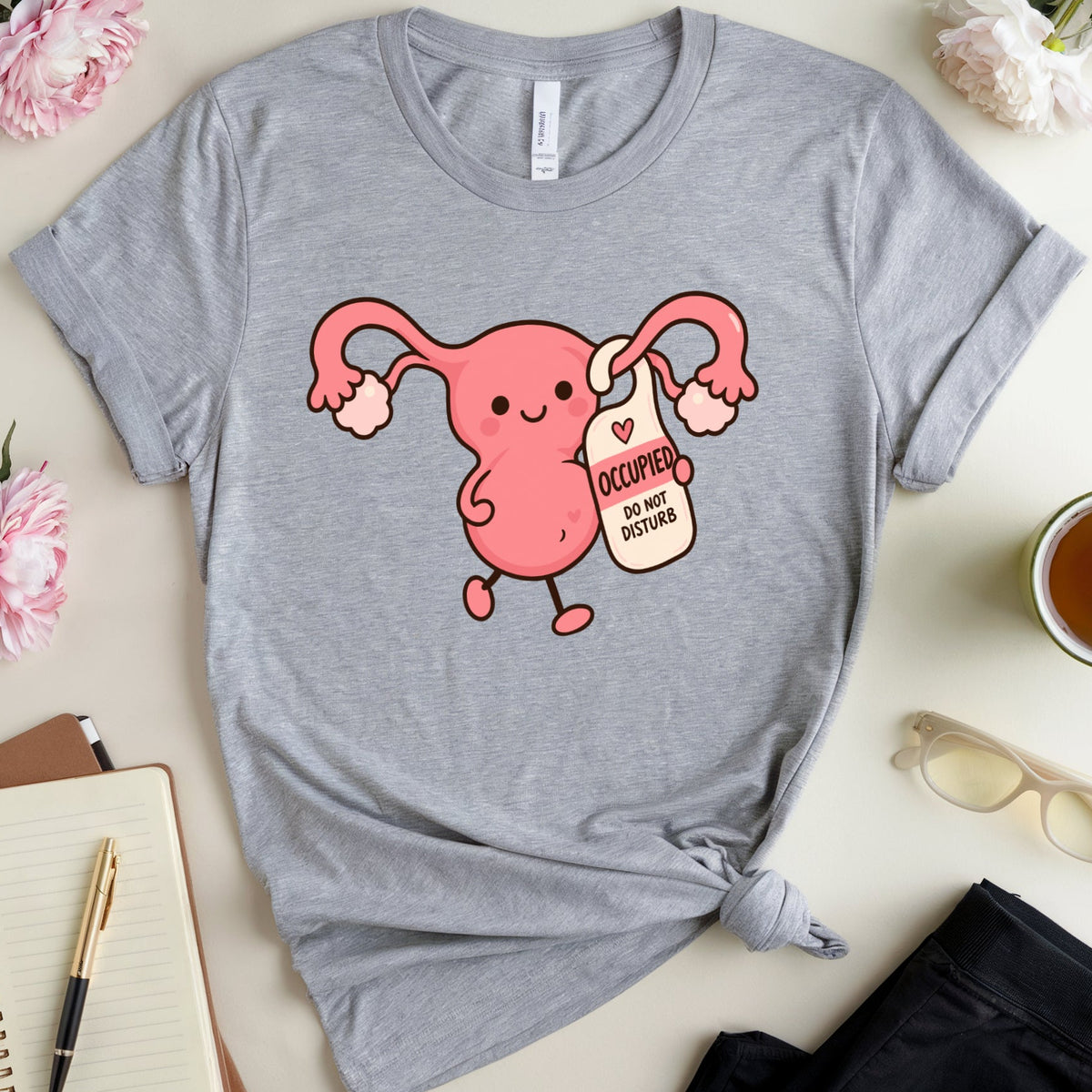 A heather gray graphic T-shirt laid flat on a light beige surface, styled with flowers, a notebook, glasses, a cup of tea, and folded black pants. The shirt features a pink cartoon uterus holding a sign that reads 'I AM OCCUPIED WITH MY OWN BODY'.