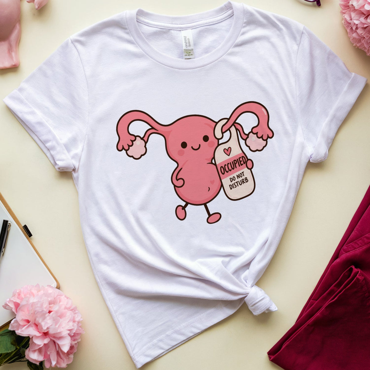 A white graphic T-shirt laid flat on a cream background, featuring a pink cartoon uterus holding a milk carton labeled 'OOCUPATION.' The shirt is styled in a flat lay with pink flowers, eyeglasses, a pink anatomical model, a notebook, and folded burgundy fabric.