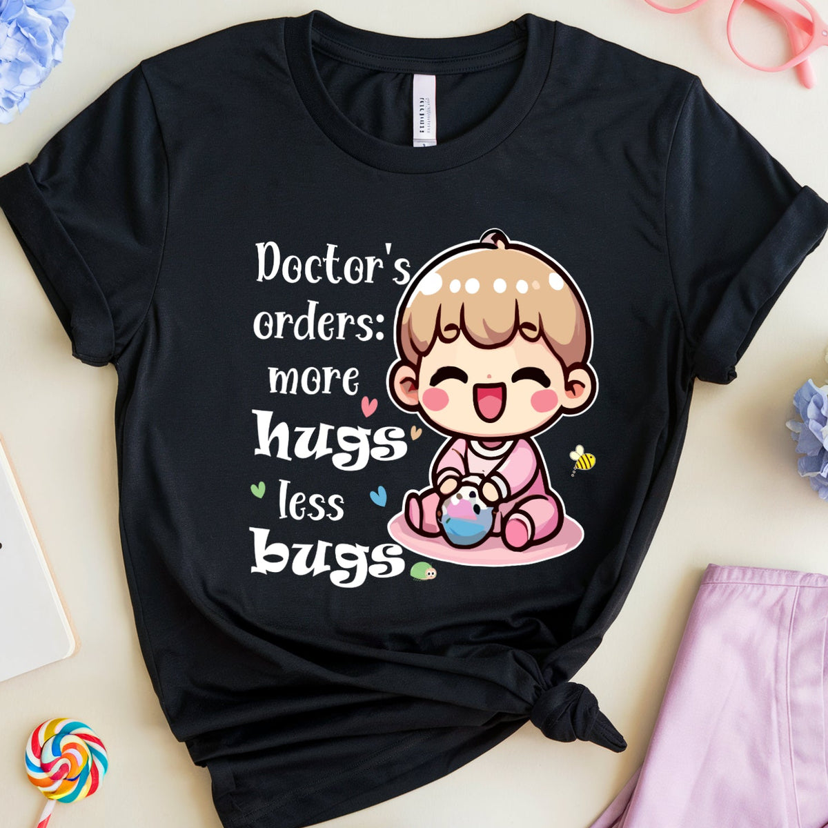 A black graphic t-shirt with a cute cartoon baby and the text 'Doctor's orders: more hugs less bugs' is displayed in a styled flat-lay arrangement with medical-themed props like pink glasses, lavender scrubs, a notebook, and flowers on a light beige background.