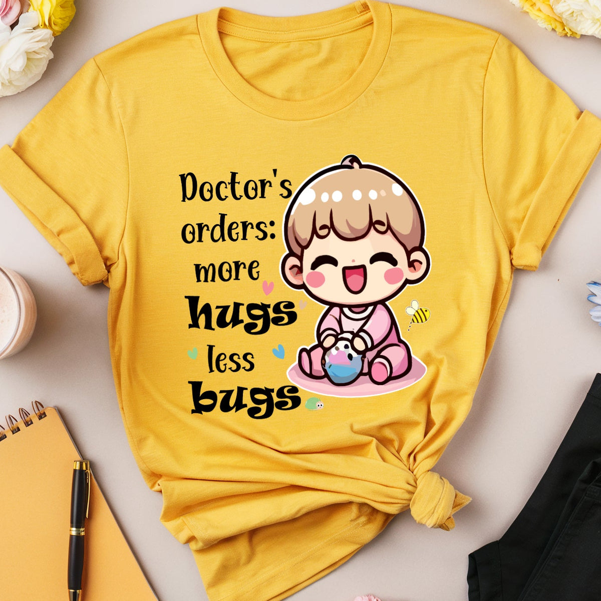 A yellow graphic T-shirt is arranged in a styled flat lay on a light beige surface. The shirt features a cartoon character and the text 'Doctor's orders: more hugs less bugs.