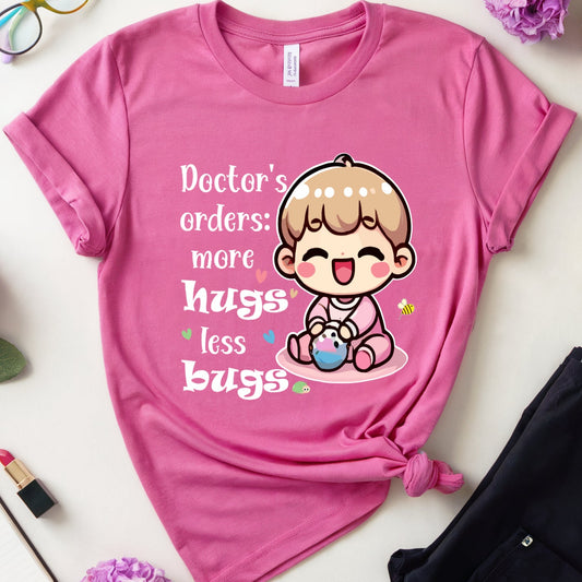 A pink graphic t-shirt with a cartoon baby and the text 'Doctor's orders: more hugs less bugs', styled in a flat lay with black pants, glasses, flowers, a notebook, and lipstick on a white background.