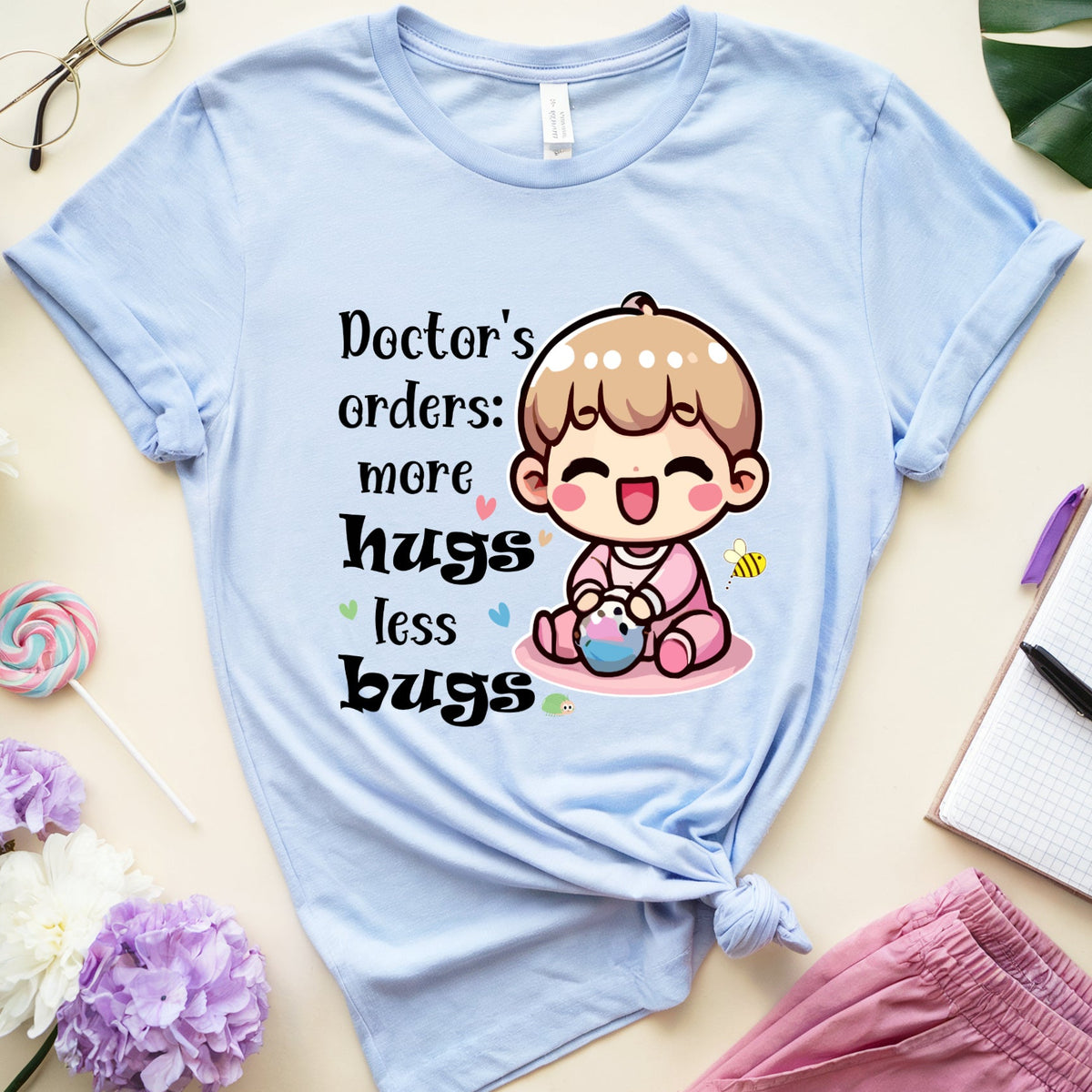 A light blue graphic t-shirt laid flat on a beige background, styled with flowers, glasses, a notebook, and pink pants. The t-shirt features a cute cartoon baby graphic and the text 'Doctor's orders: more hugs less bugs'.