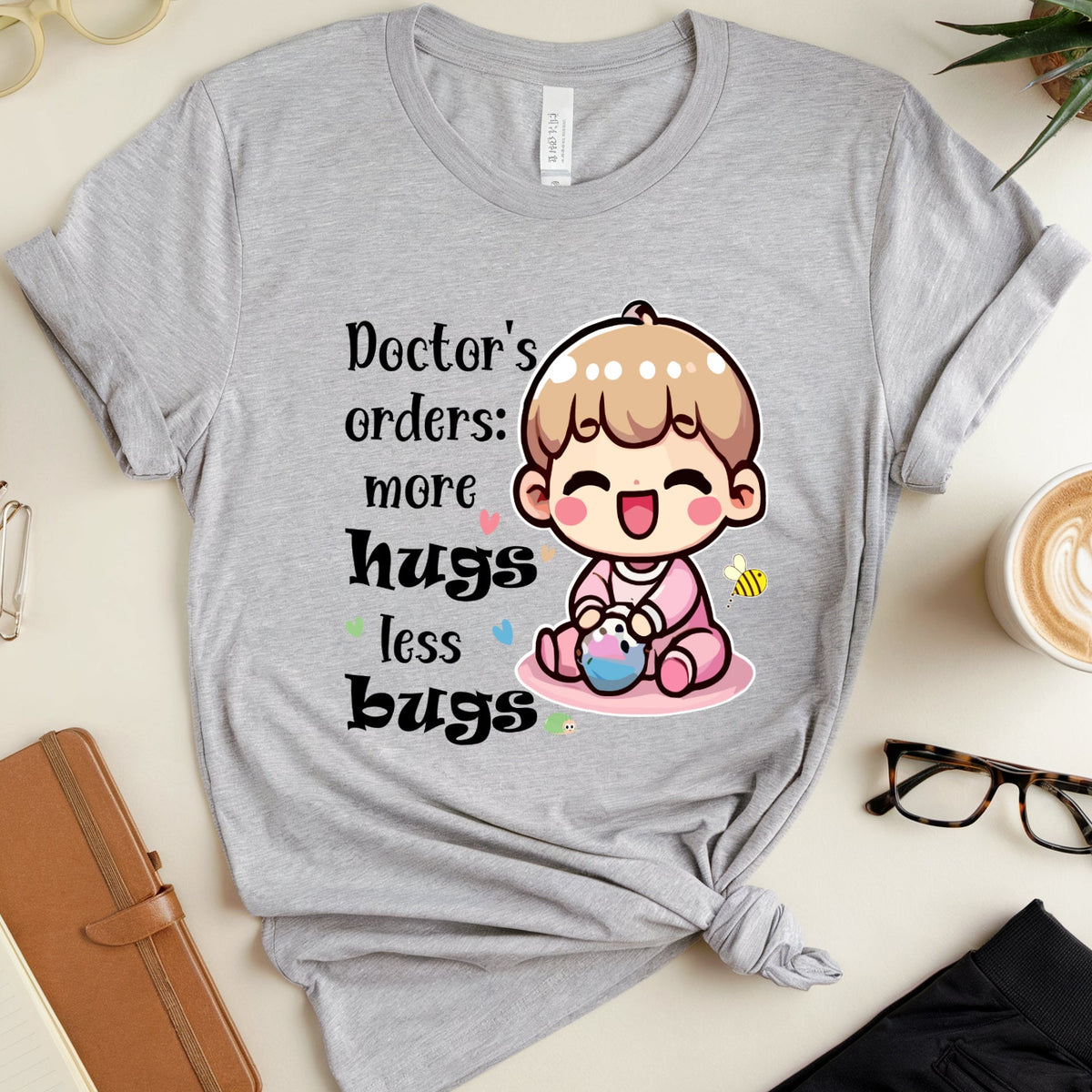 A heather gray graphic t-shirt with a cute cartoon baby design and the text 'Doctor's orders: more hugs less bugs', styled in a flat lay with eyeglasses, a notebook, a plant, and a cup of latte.