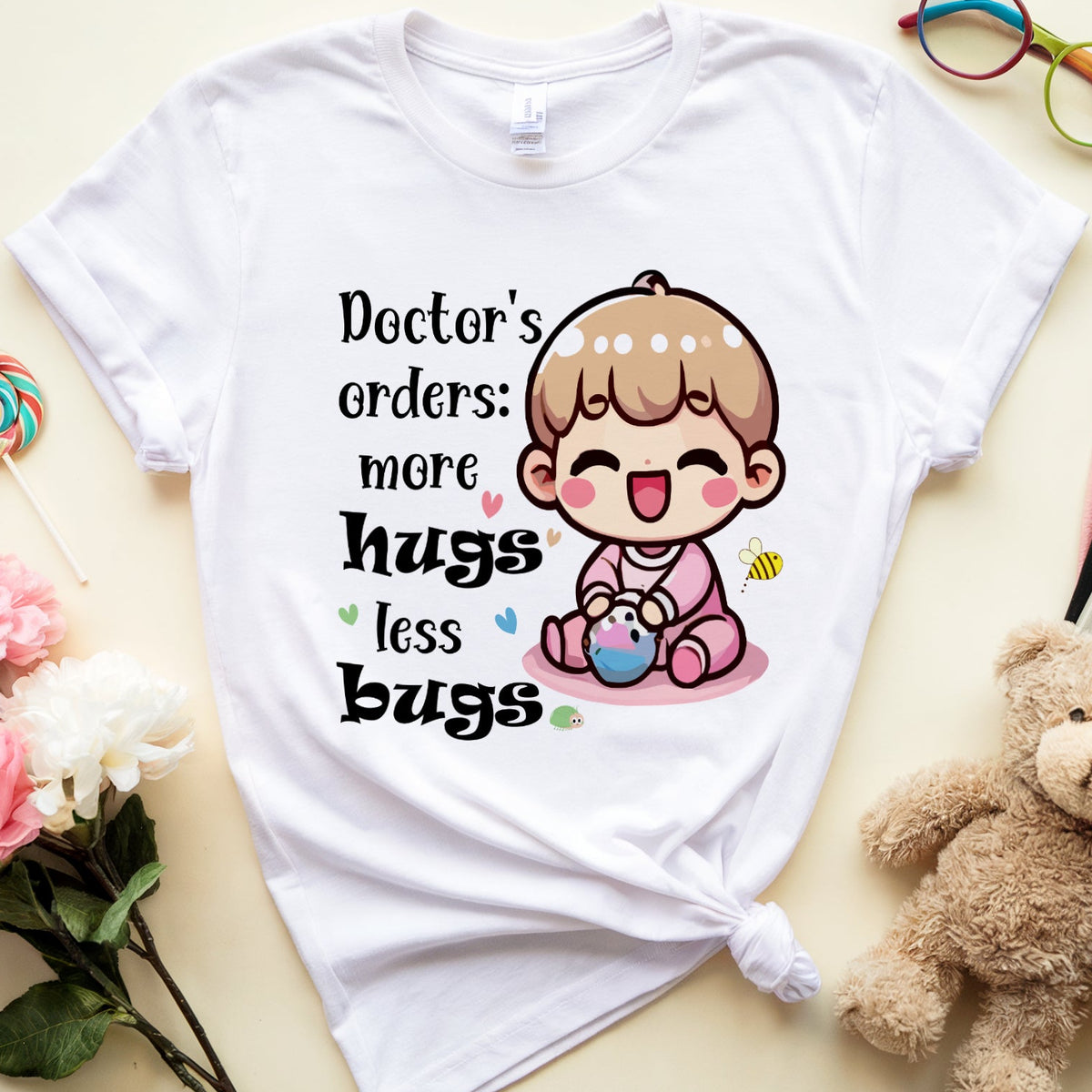 A white graphic t-shirt with a cute cartoon baby and the text 'Doctor's orders: more hugs less bugs', styled in a flat lay with a teddy bear, flowers, glasses, and a book on a light beige background.