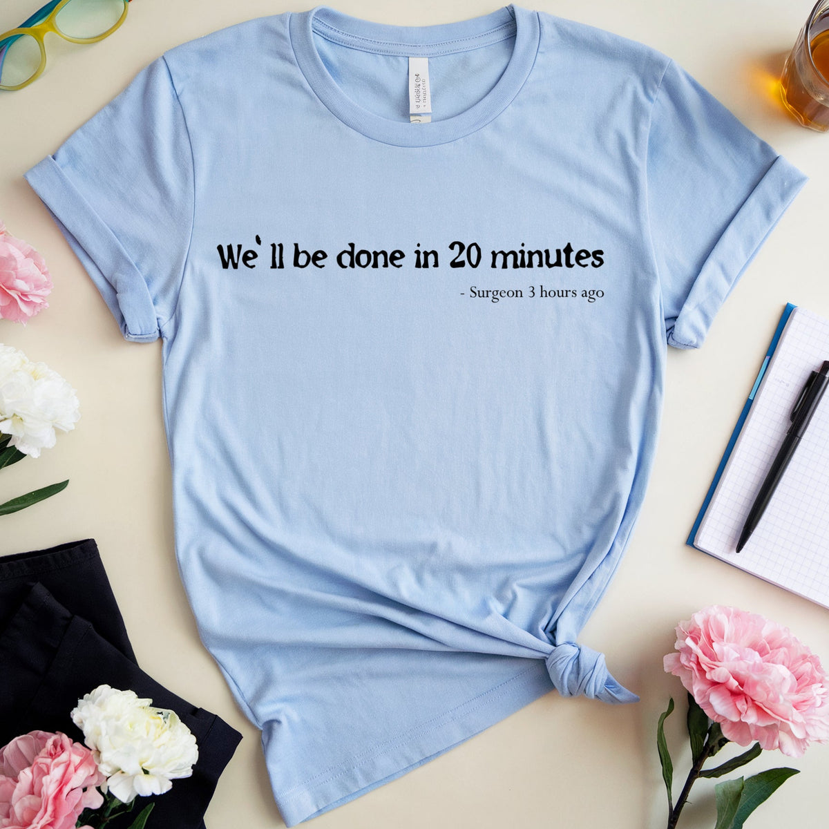 A light blue graphic t-shirt with a black printed quote, styled in a flat lay with flowers, glasses, a notebook, and a drink on a beige background.