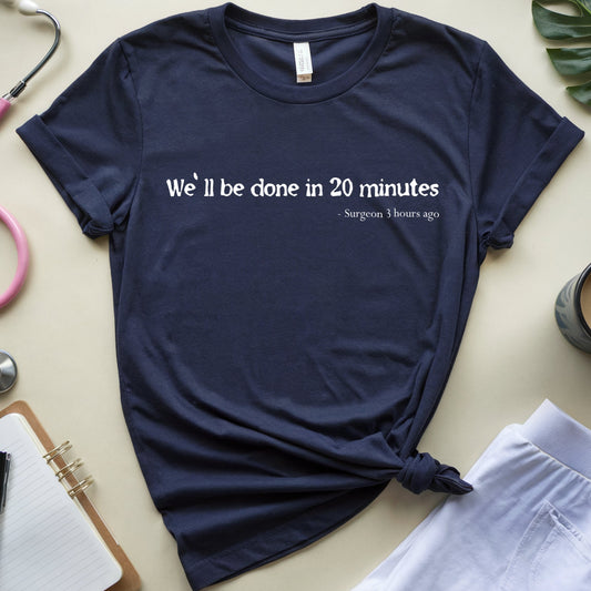A navy blue graphic t-shirt with a white printed quote by Hippocrates, styled in a flat lay with a pink stethoscope, notebook, coffee cup, and green leaves on a beige background.