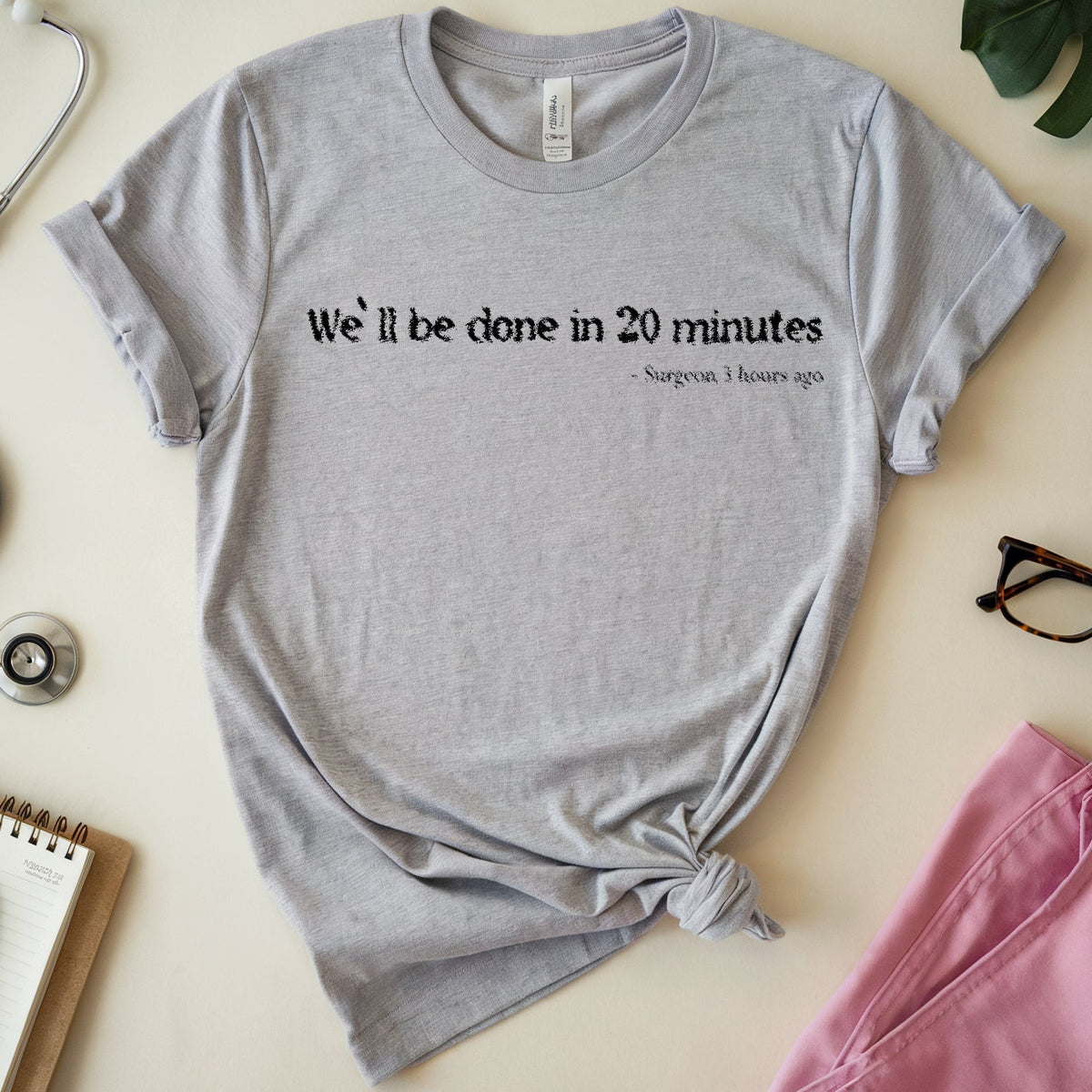 A heather gray short-sleeve T-shirt with rolled cuffs and a tied hem, featuring the printed text 'We'll be done in 20 minutes' and a smaller subtitle. The shirt is styled in a flat lay on a light beige surface with a stethoscope, notebook, pen, eyeglasses, green leaves, and pink fabric around it.
