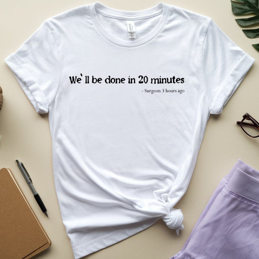 A white graphic t-shirt with the text 'We'll be clone in 20 minutes - Surgeon 3 hours ago' printed on the chest, styled in a flat-lay arrangement with a notebook, glasses, a cup, and other office accessories on a beige background.