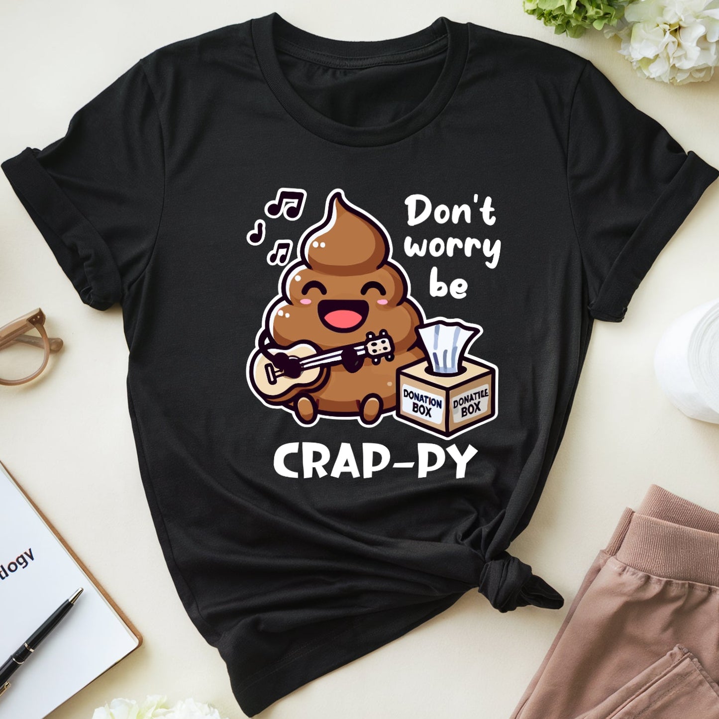 Black t-shirt with a humorous design featuring poop singing don't worry be crappy with tissue donation box.