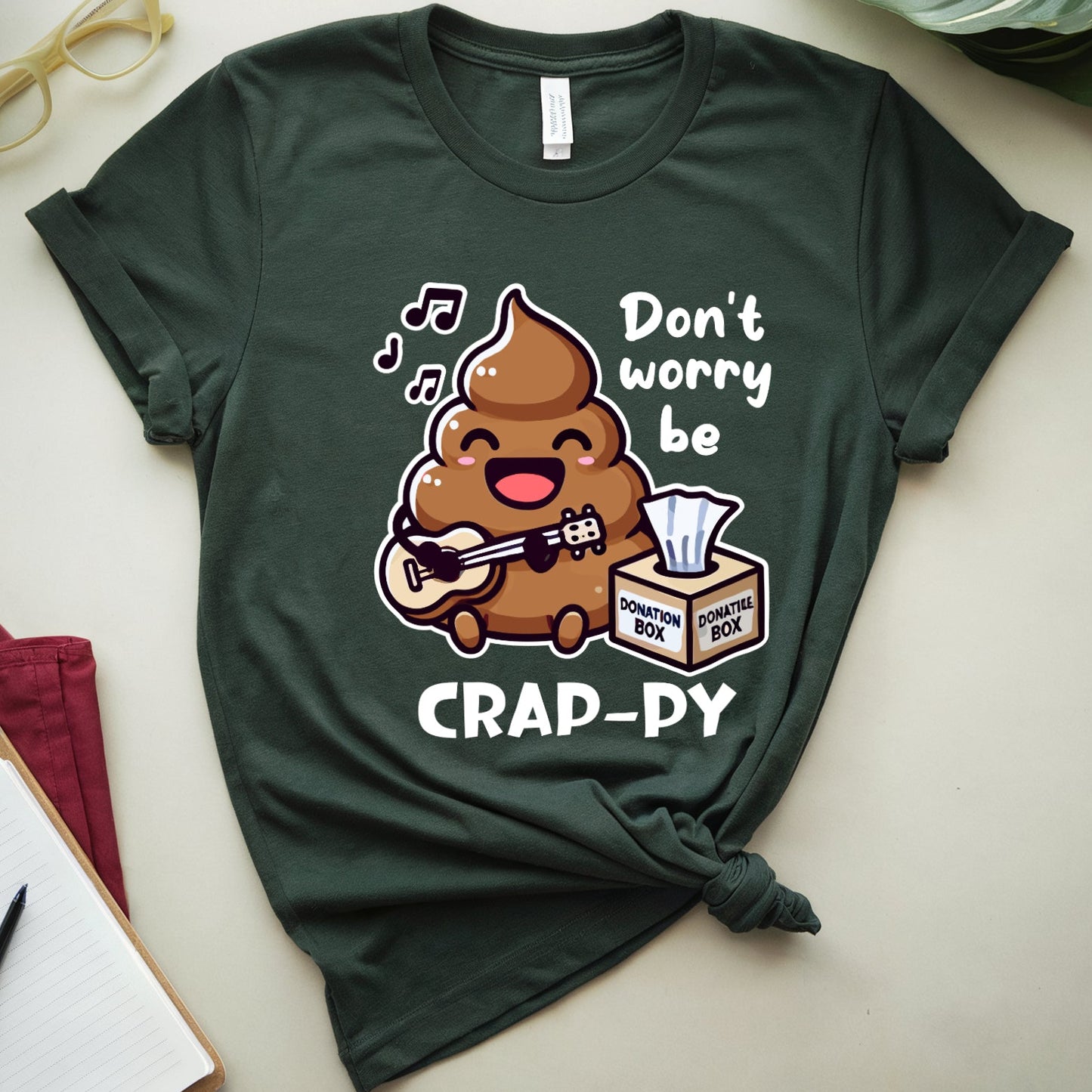 Green t-shirt with a cartoon poop singing don't worry be crappy with tissue donation box.