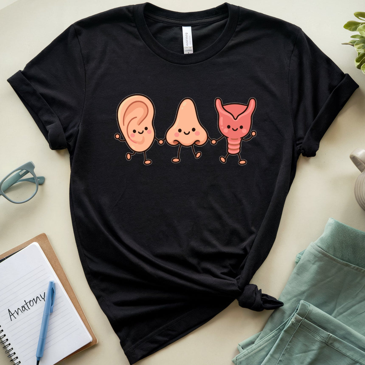A black short-sleeve graphic T-shirt laid flat on a light beige surface, styled with a knot at the hem. The shirt features a cartoon anatomical illustration of an ear, nose, and throat in peach and coral tones.