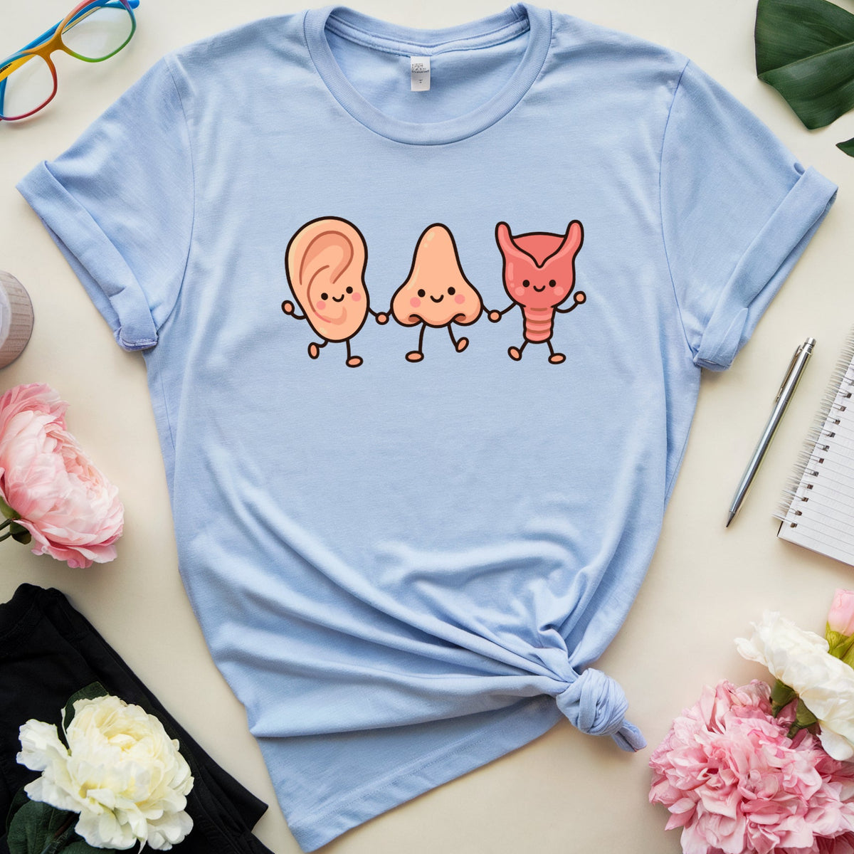 A light blue graphic t-shirt with a cartoon ear, nose, and throat illustration on the chest, styled in a flat lay with flowers, glasses, a notebook, and a pen on a beige background.