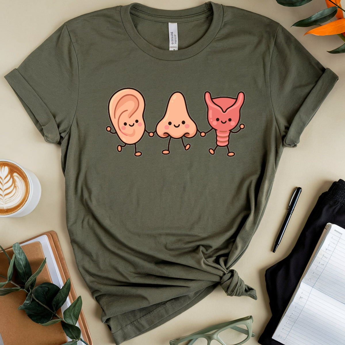 An olive green graphic t-shirt is displayed in a styled flat-lay arrangement on a beige background. The t-shirt features a cartoon graphic of three anthropomorphic body parts: an ear, a stomach, and a bow.