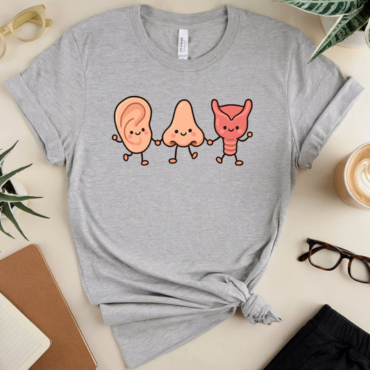 A heather gray short-sleeve T-shirt laid flat on a light beige surface, styled with glasses, plants, a notebook, a coffee cup, and black shorts. The shirt has a centered graphic of three cute cartoon body parts: a peach ear, a pale pink nose, and a coral throat, all holding hands.