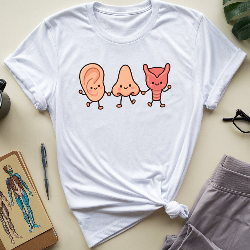 A white short-sleeve T-shirt laid flat on a light beige surface, styled with a knot at the bottom. The shirt has a cartoon graphic of three smiling body parts holding hands: a peach-colored ear, a pinkish triangle shape, and a red horn-like form.