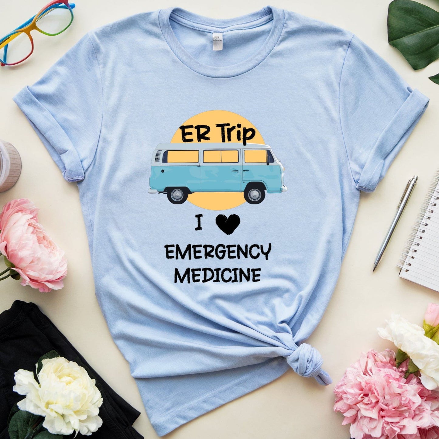 Light blue t-shirt with 'ER Trip I ❤️ Emergency Medicine' graphic on a light surface with flowers and glasses.