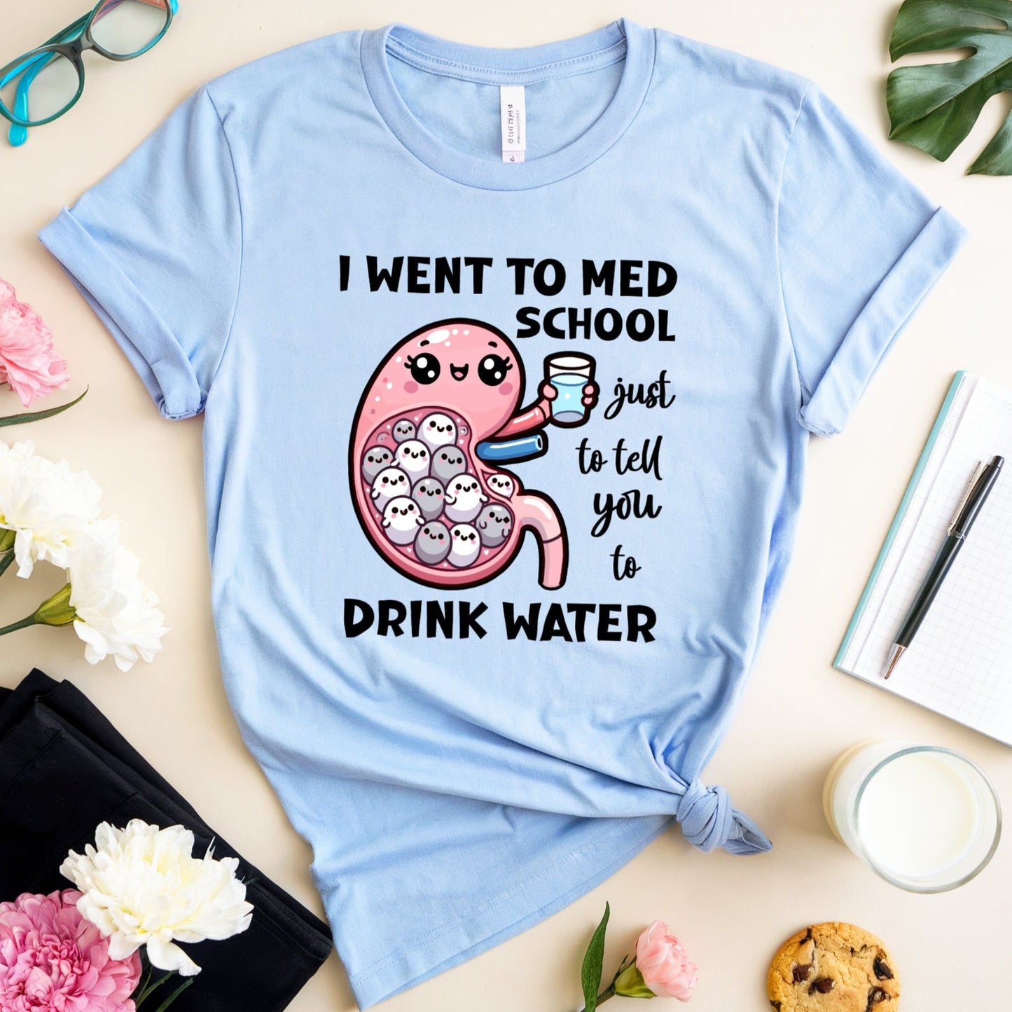 Light blue t-shirt with a graphic design and text on a table with flowers and a glass of milk.