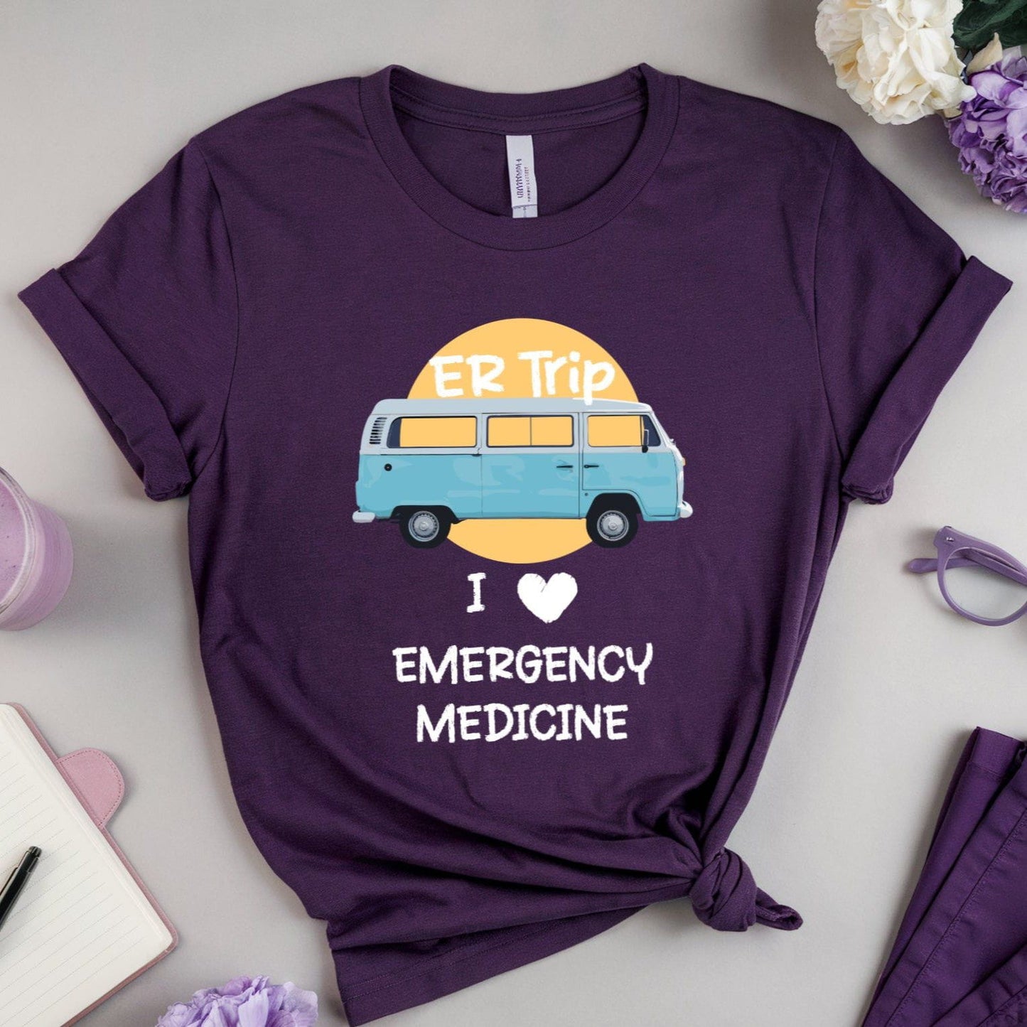Purple t-shirt with a graphic of a van and text 'I love emergency medicine' on a light gray surface with flowers and a drink.
