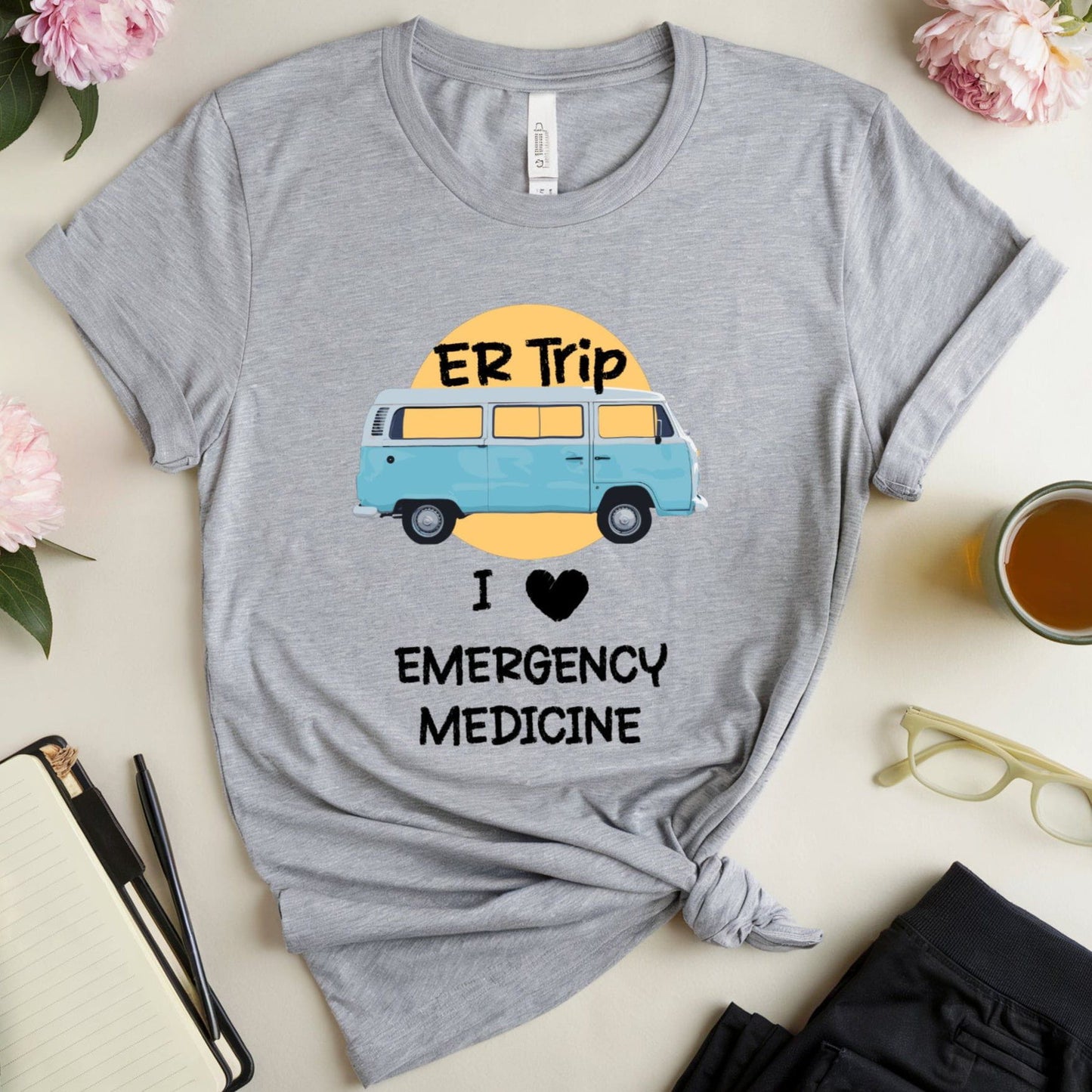 Gray t-shirt with a graphic of a van and text 'ER Trip I ❤️ Emergency Medicine' on a light surface with flowers and a mug.