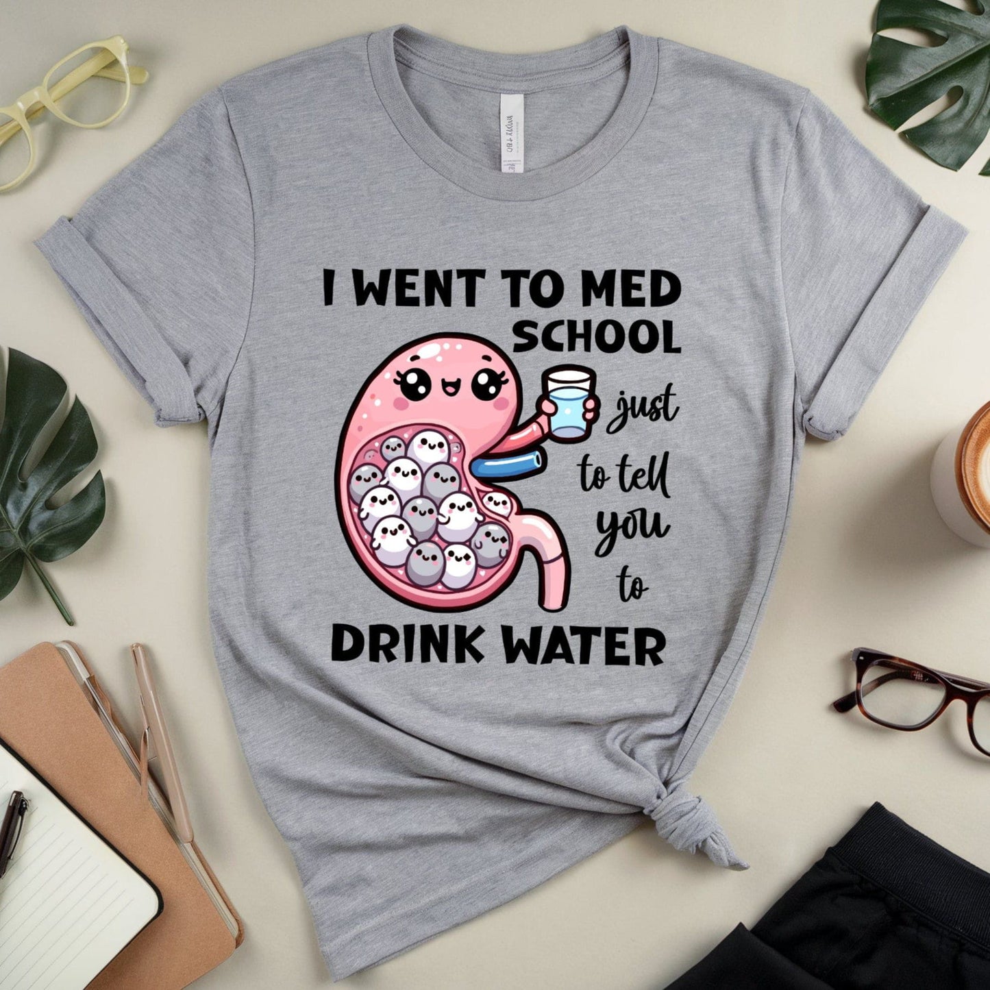 Gray t-shirt with a humorous design about going to med school, displayed on a surface with accessories.