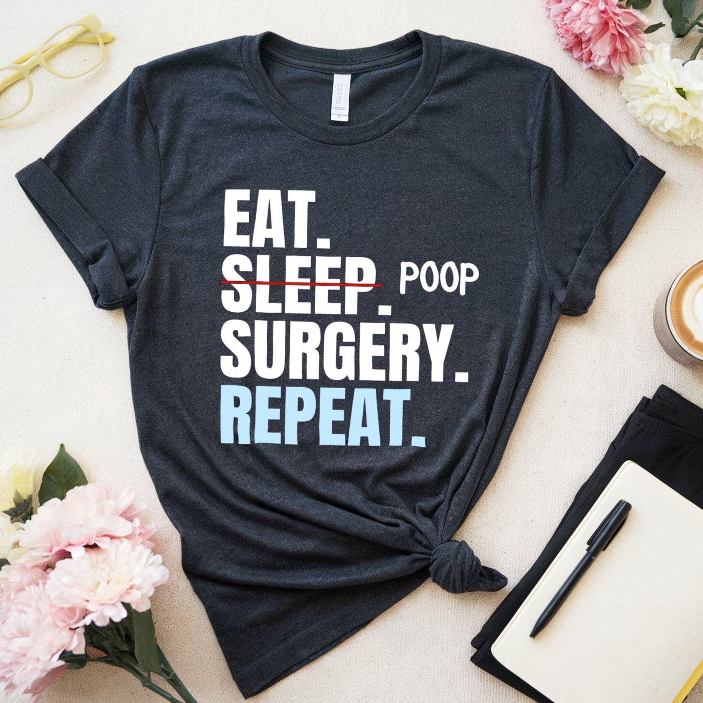 Black t-shirt with 'EAT. SLEEP. POOP. SURGERY. REPEAT.' text on a light surface with flowers and a cup of coffee.