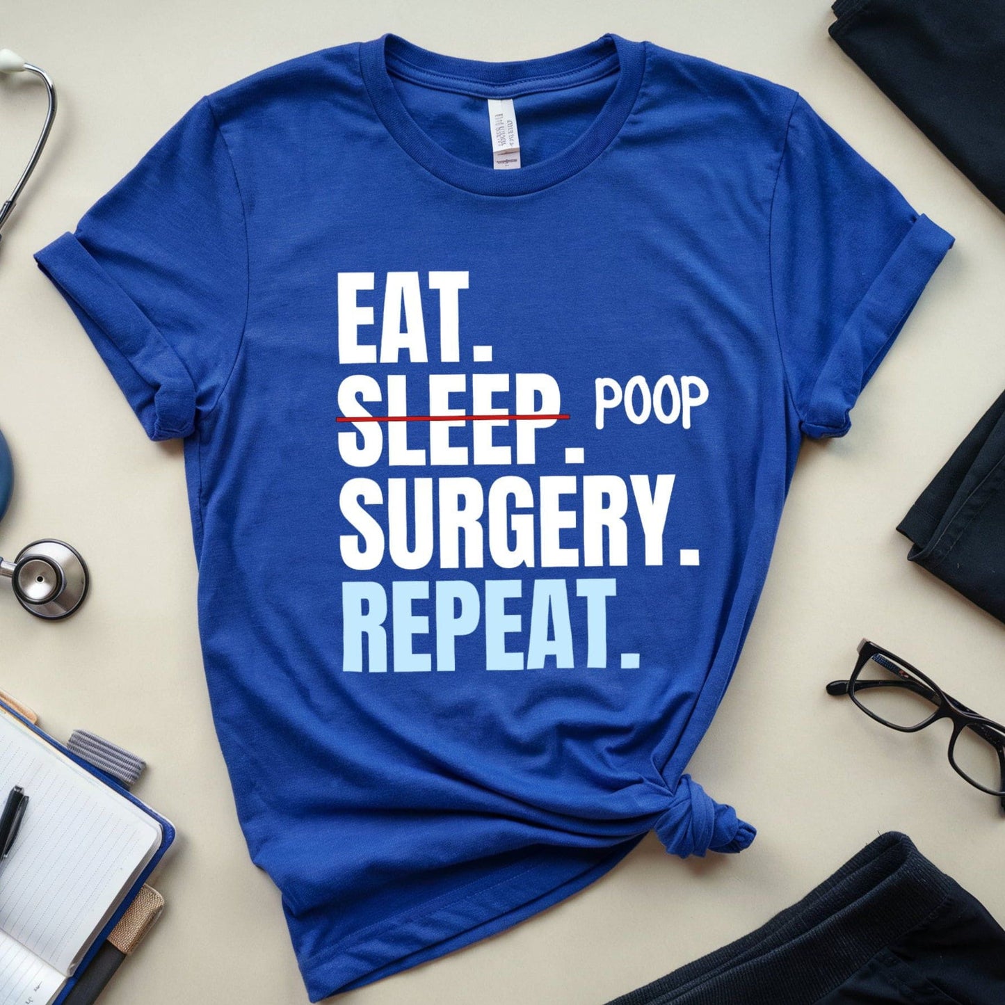 Blue t-shirt for surgeons with 'Eat. Sleep. Poop. Surgery. Repeat.' text on a light surface with medical-themed items.