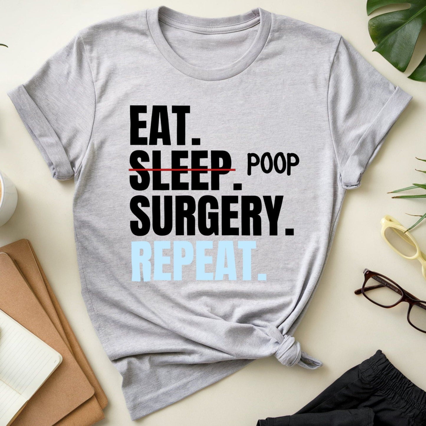 Gray t-shirt for surgeons with 'Eat. Sleep. Poop. Surgery. Repeat.' text on a light surface with office items.