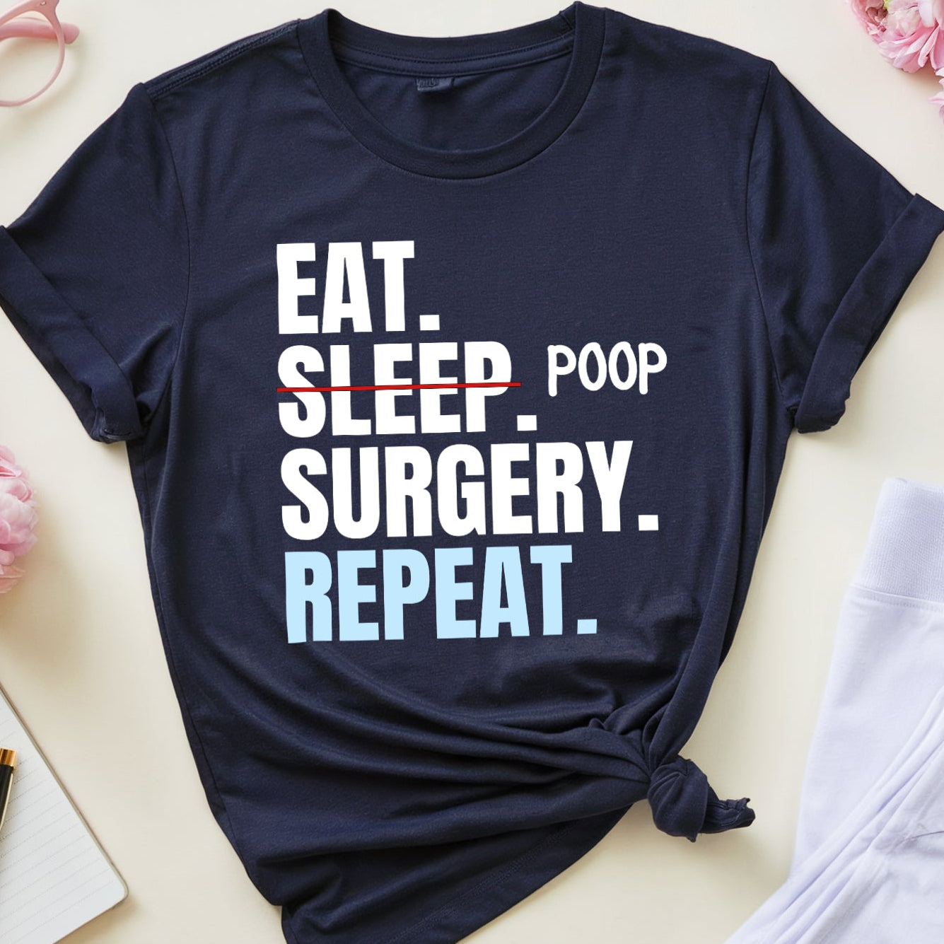 Navy blue t-shirt with 'Eat. Sleep. Poop. Surgery. Repeat.' text on a light surface with flowers and glasses.