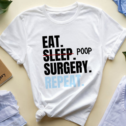 White t-shirt with 'Eat. Sleep. Poop. Surgery. Repeat.' text on a beige surface with accessories.