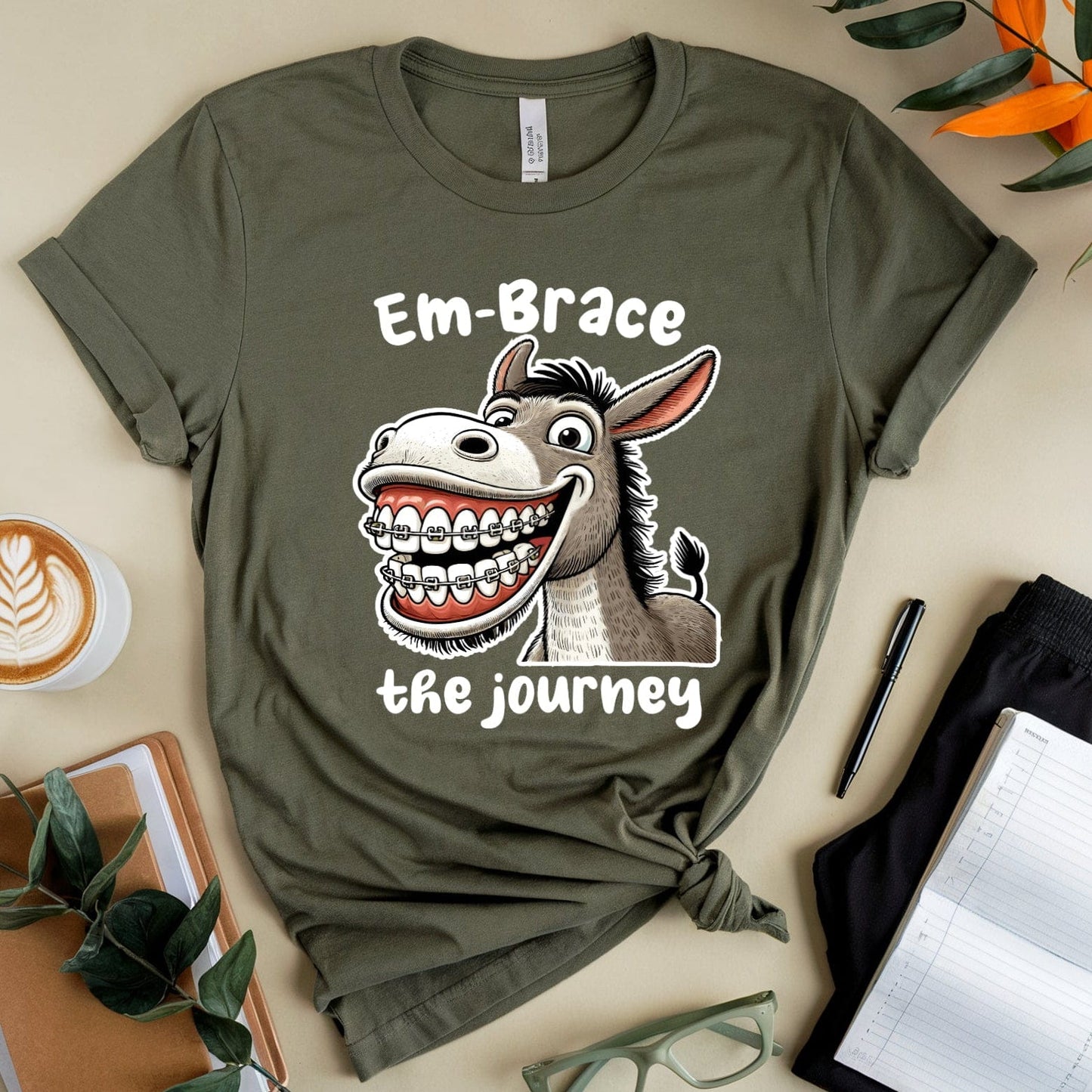 Green t-shirt with a donkey graphic and text on a beige surface with a cup of coffee, notebook, and glasses.
