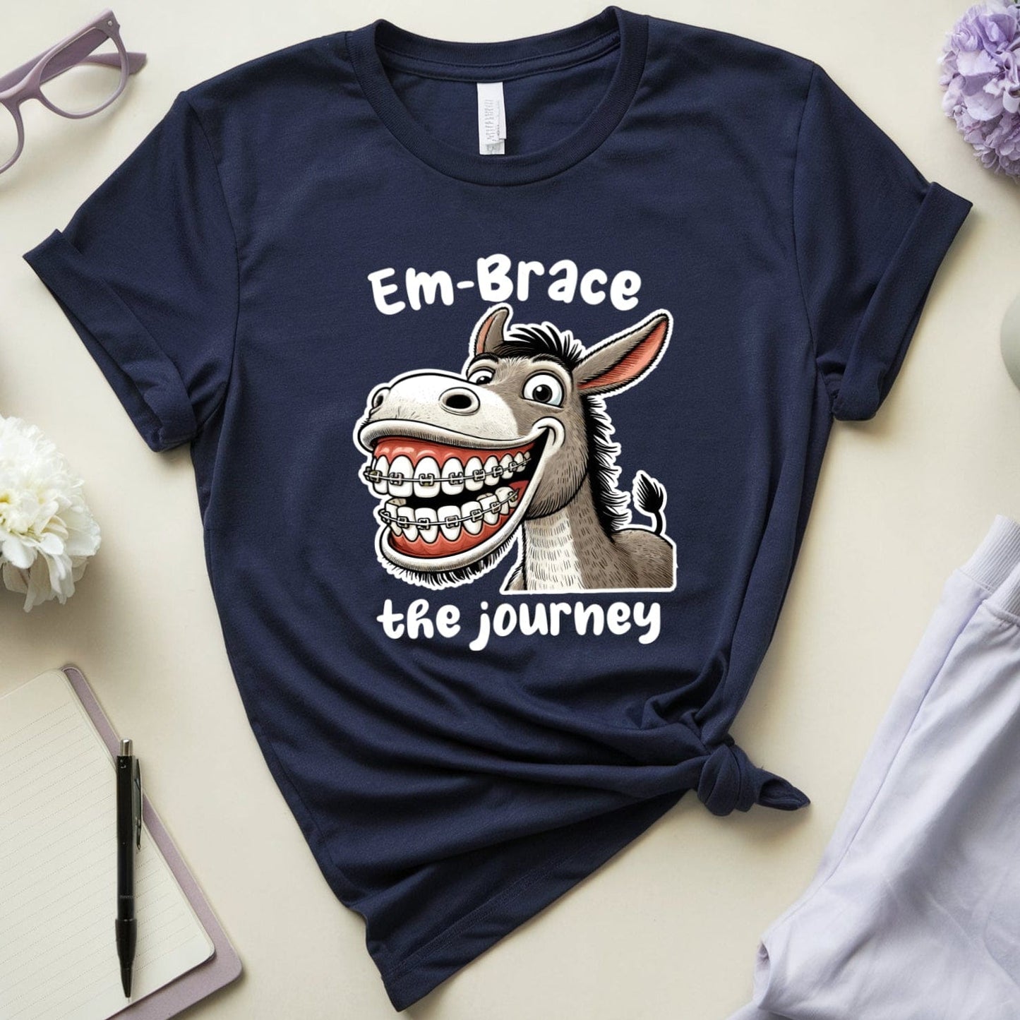 Navy blue t-shirt with a donkey graphic and text 'Em-Brace the journey' on a light surface with accessories.