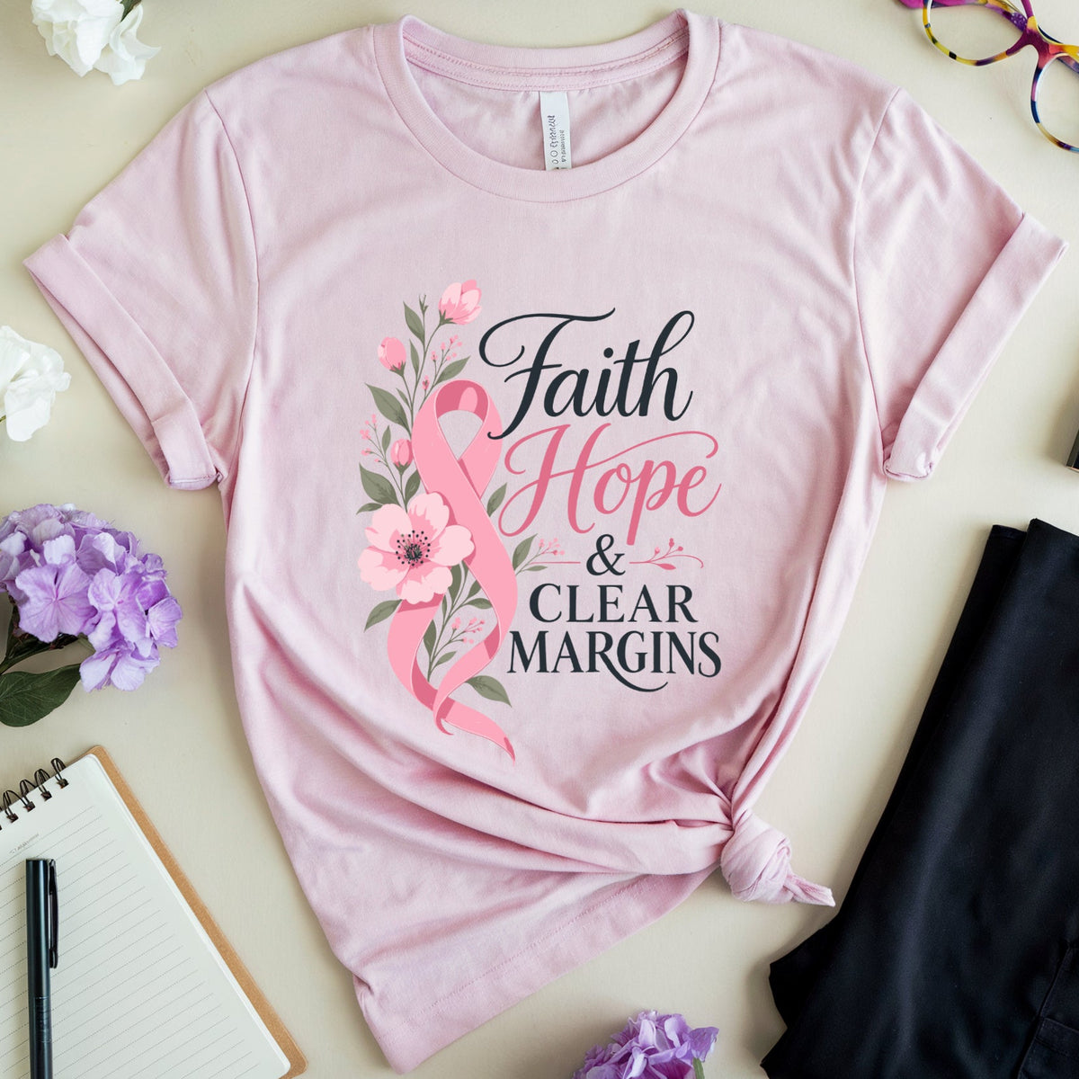 A light pink graphic T-shirt laid flat on a beige surface, styled with flowers, glasses, a notebook, pen, lipstick, and folded dark pants. The shirt has a floral and ribbon design with the text 'Faith Hope & Clear Margins.