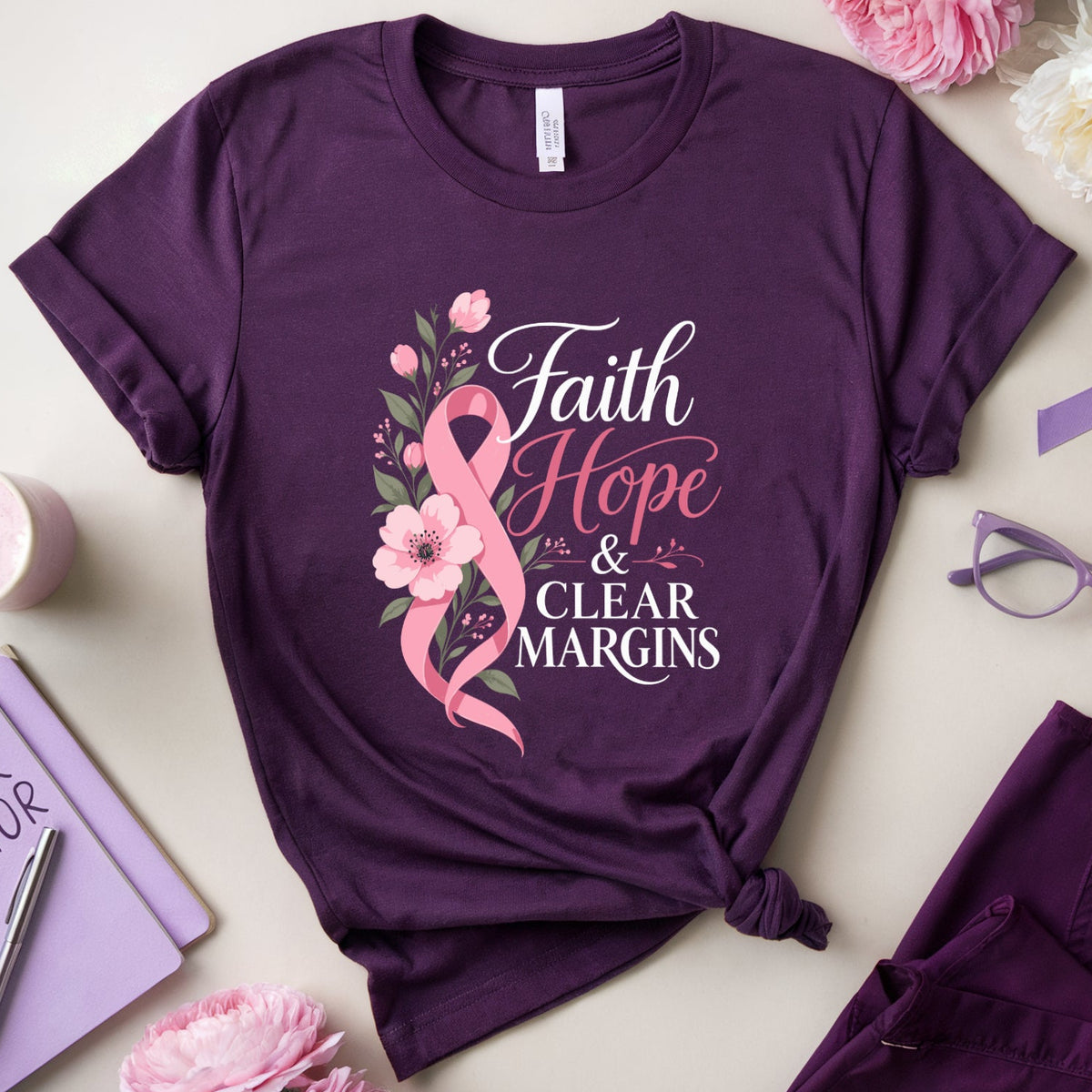 A styled flat-lay of a purple short-sleeve T-shirt with a pink and white floral graphic reading 'Faith Hope & Clear Margins,' surrounded by pink and white flowers, a purple awareness ribbon, purple eyeglasses, a purple notebook, and matching purple scrubs on a light beige background.