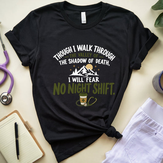 A black graphic t-shirt with a medical-themed quote printed on the front, styled in a flat lay with a stethoscope, notebook, coffee mug, and white pants on a light beige background.