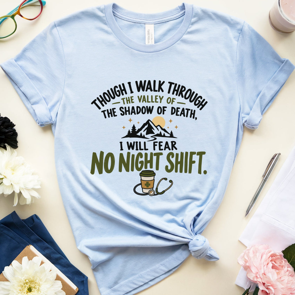 A light blue graphic t-shirt with a 'No Night Shift' design, styled in a flat lay with glasses, flowers, a drink, and stationery on a light beige background.
