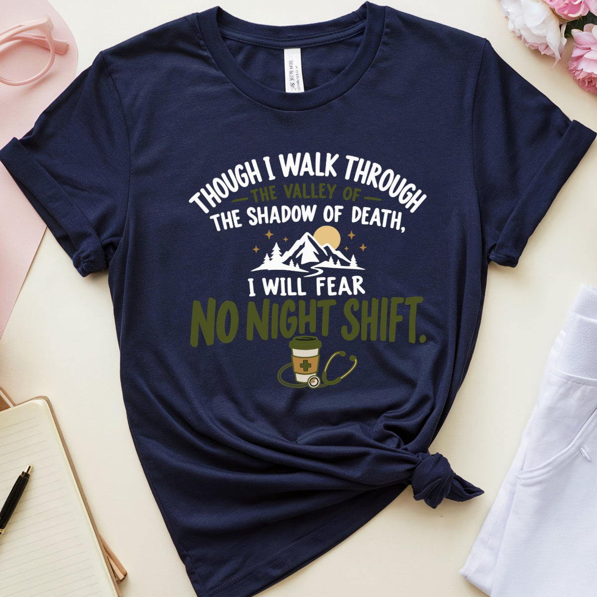 A navy blue graphic t-shirt with a motivational quote about night shifts, styled in a flat lay with pink flowers, a notebook, glasses, a mug, and white pants on a light beige background.