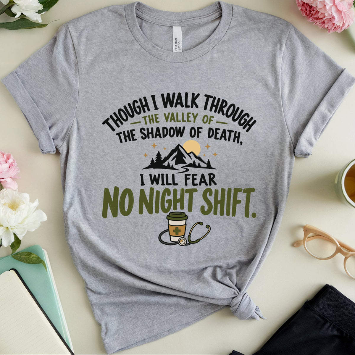 A heather gray graphic t-shirt with a 'no night shift' theme, styled in a flat-lay arrangement with flowers, glasses, a notebook, and a cup of tea on a light beige background.