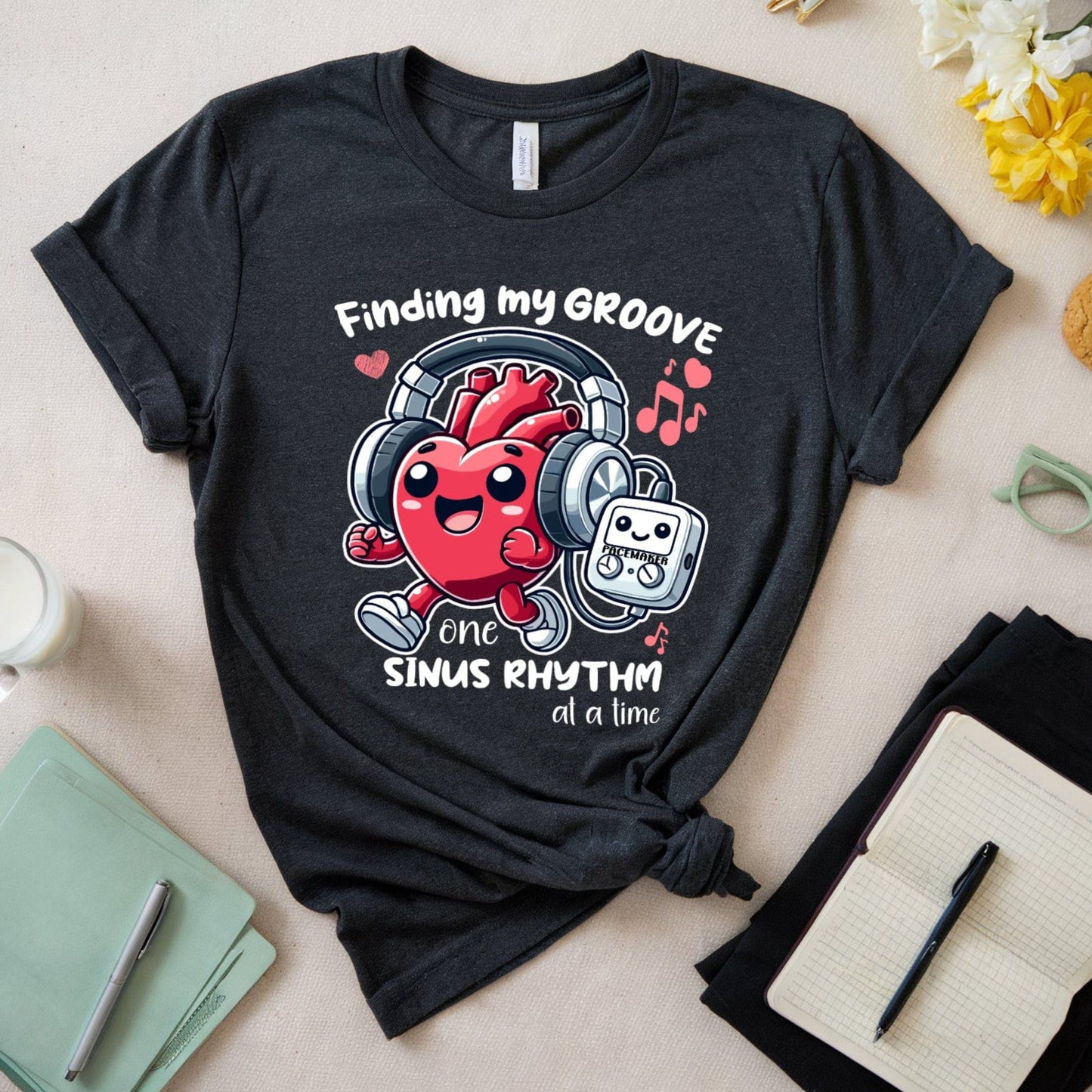 Black t-shirt  for cardiology with a jogging heart listening to music from its pacemaker and text 'Finding my GROOVE one sinus RHYTHM at a time' on a light surface with accessories.