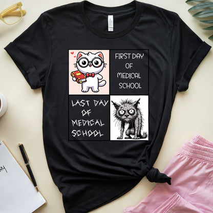 A black graphic t-shirt featuring a cartoon cat illustration and the text 'FIRST DAY OF MEDICAL SCHOOL' and 'LAST DAY OF MEDICAL SCHOOL' in a two-panel design. The t-shirt is styled in a flat lay with pink pants, a coffee cup, a notebook, and glasses on a light beige background.