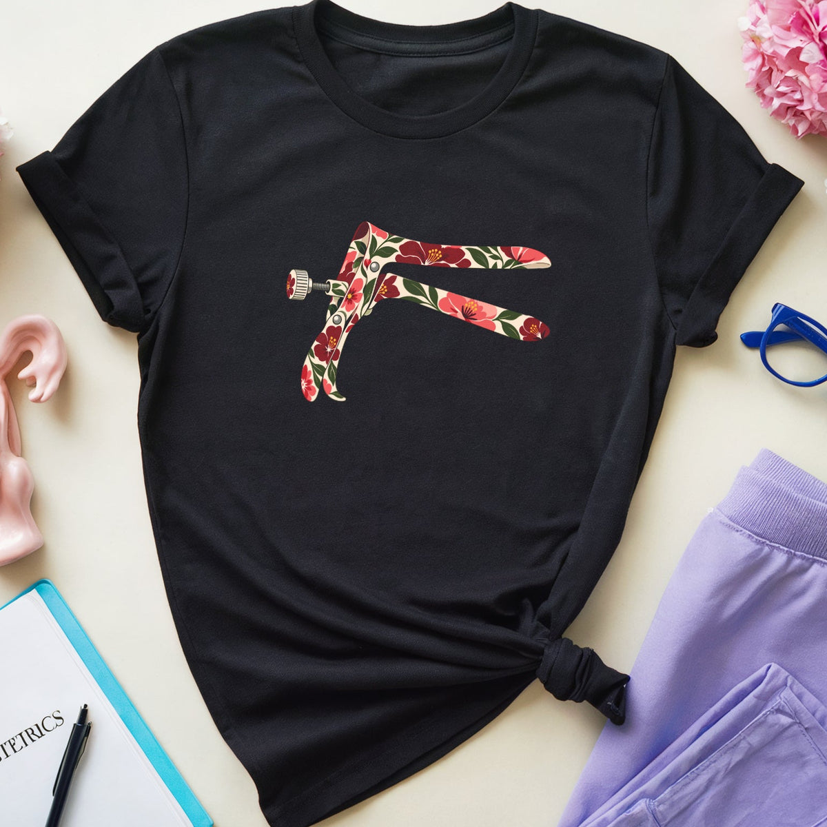 A black short-sleeve T-shirt with a colorful floral graphic of crossed legs, styled in a flat lay with lavender sweatpants, pink flowers, blue glasses, a pink anatomical model, and a notebook on a light beige background.