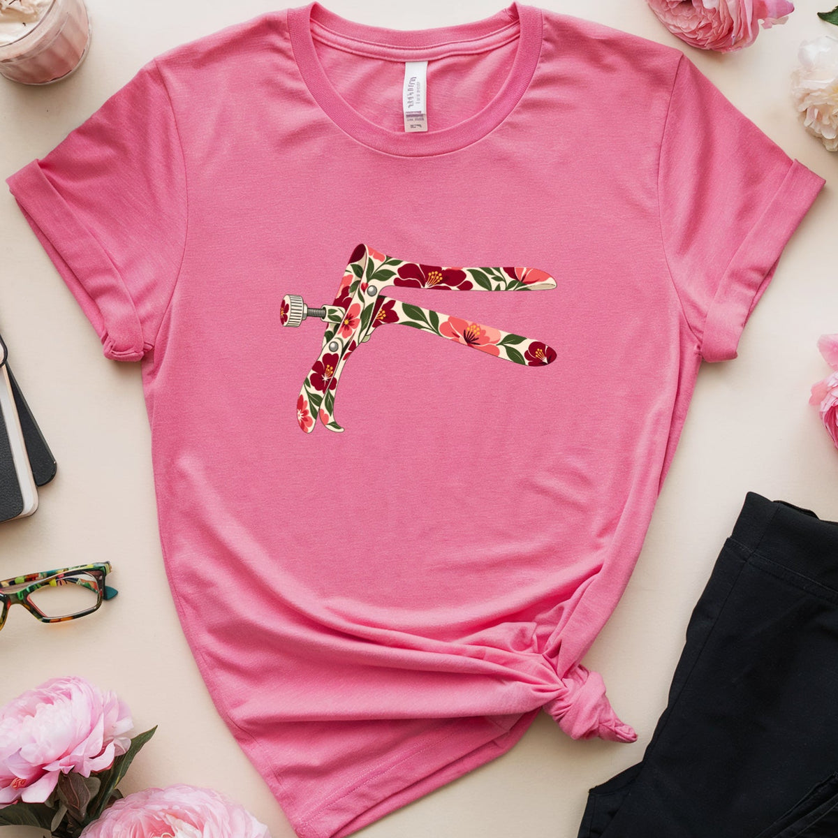 A pink short-sleeve t-shirt with a colorful floral-patterned dragonfly graphic on the chest, styled in a flat lay with black pants, pink flowers, glasses, a notebook, and a drink on a light beige background.