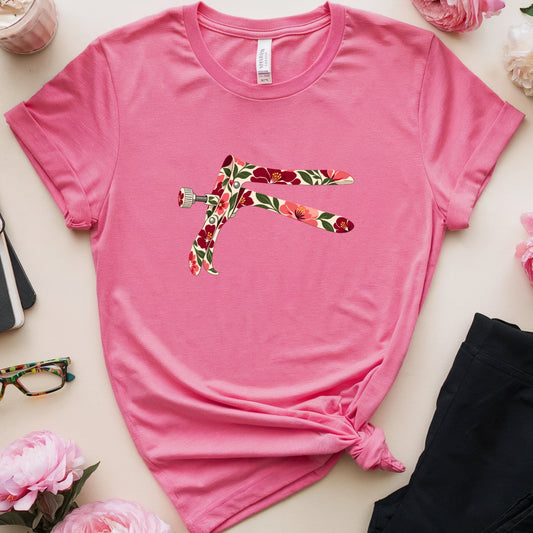 A pink short-sleeve t-shirt with a colorful floral-patterned dragonfly graphic on the chest, styled in a flat lay with black pants, pink flowers, glasses, a notebook, and a drink on a light beige background.