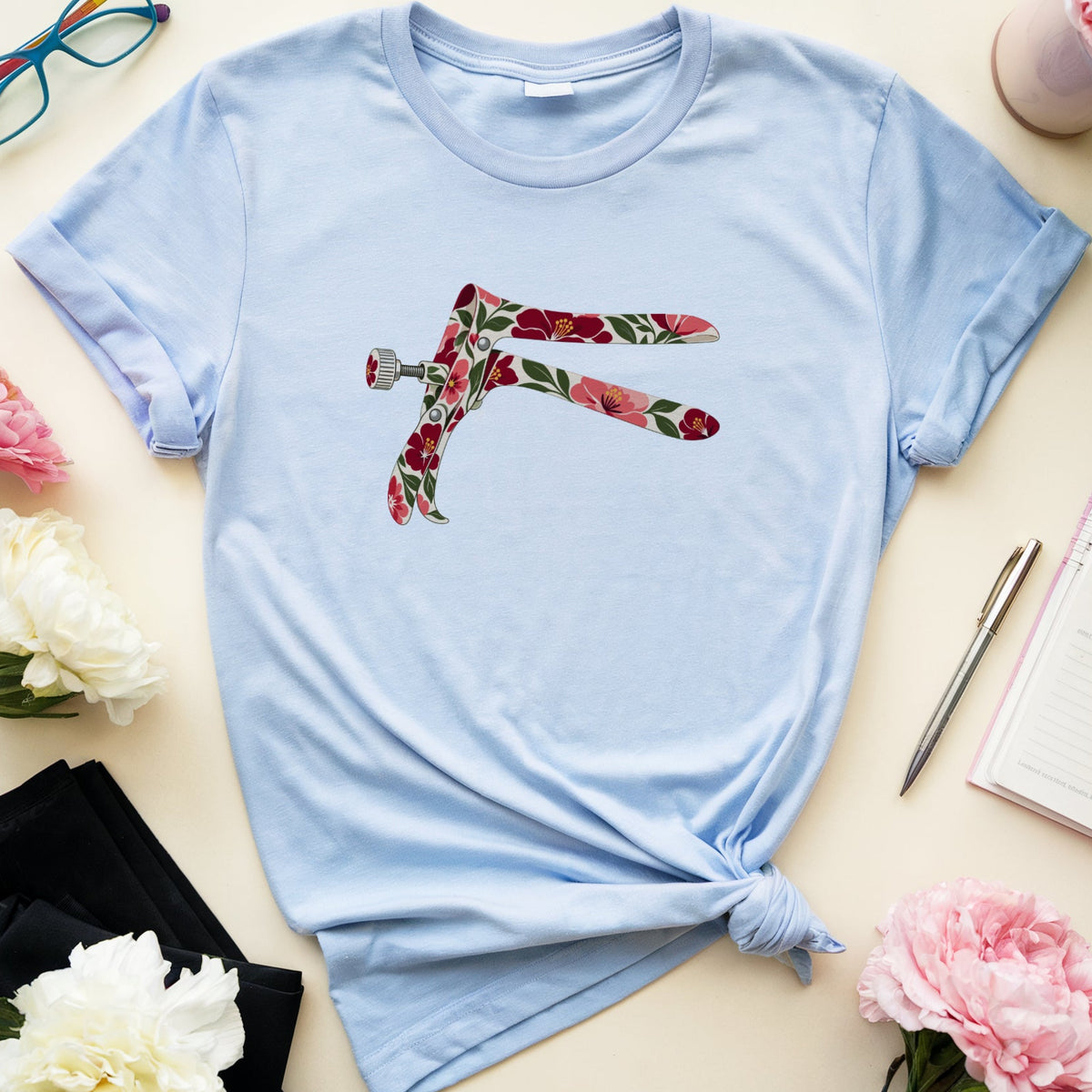 A light blue short-sleeve t-shirt with a floral-patterned dagger graphic on the chest, styled in a flat lay with pink and white flowers, glasses, a notebook, and a pen on a cream-colored background.