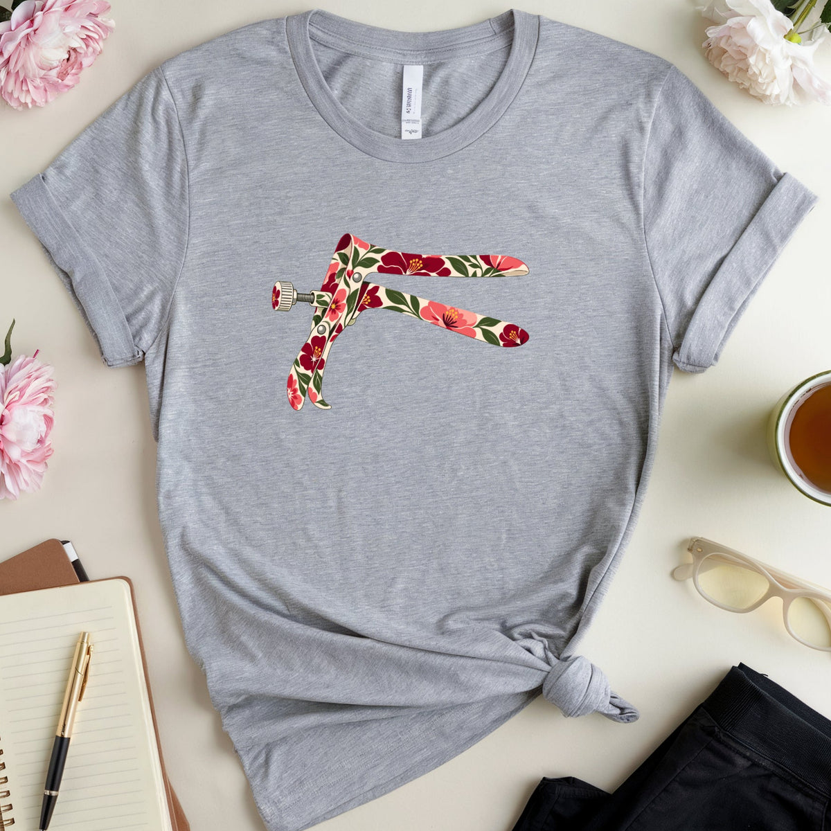 A heather gray short-sleeve t-shirt with a colorful floral 'F' graphic on the chest, styled in a flat lay with pink flowers, a notebook, glasses, and a cup of tea on a light beige background.