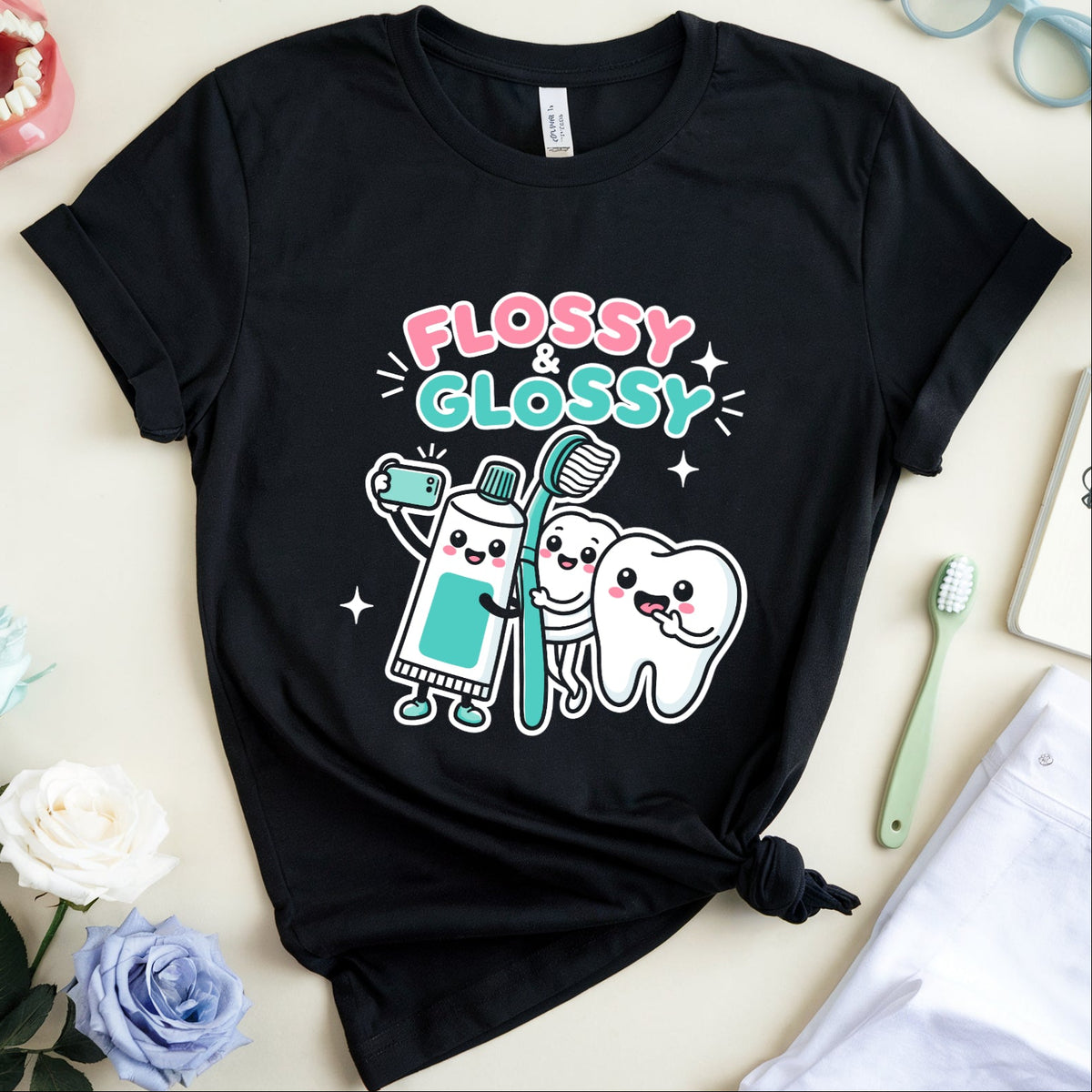 A black graphic t-shirt featuring cartoon characters of a toothbrush and a tooth with the text 'FLOSSY & GLOSSY' in pink and teal. The shirt is styled in a flat lay with dental-themed props including a model mouth, glasses, a notebook, a toothbrush, and flowers on a light beige background.