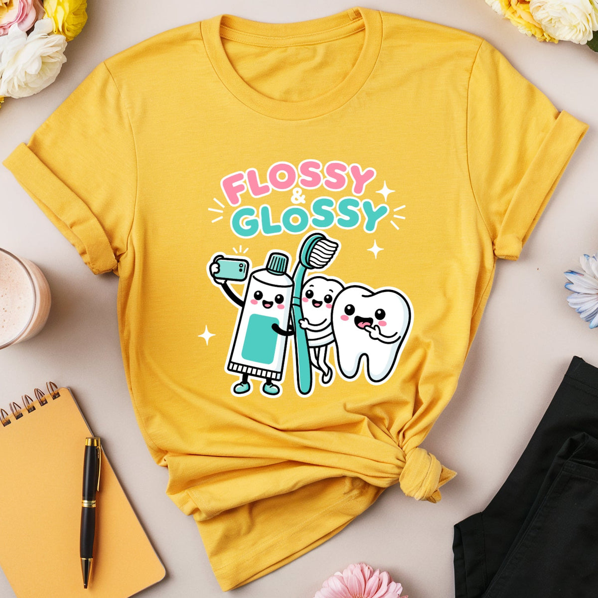 A yellow graphic t-shirt with a cute cartoon toothbrush and tooth design that reads 'FLOSSY GLOSSY' in pink and blue. The t-shirt is styled in a flat lay with black pants, a yellow notebook, a pink drink, and flowers on a light beige background.